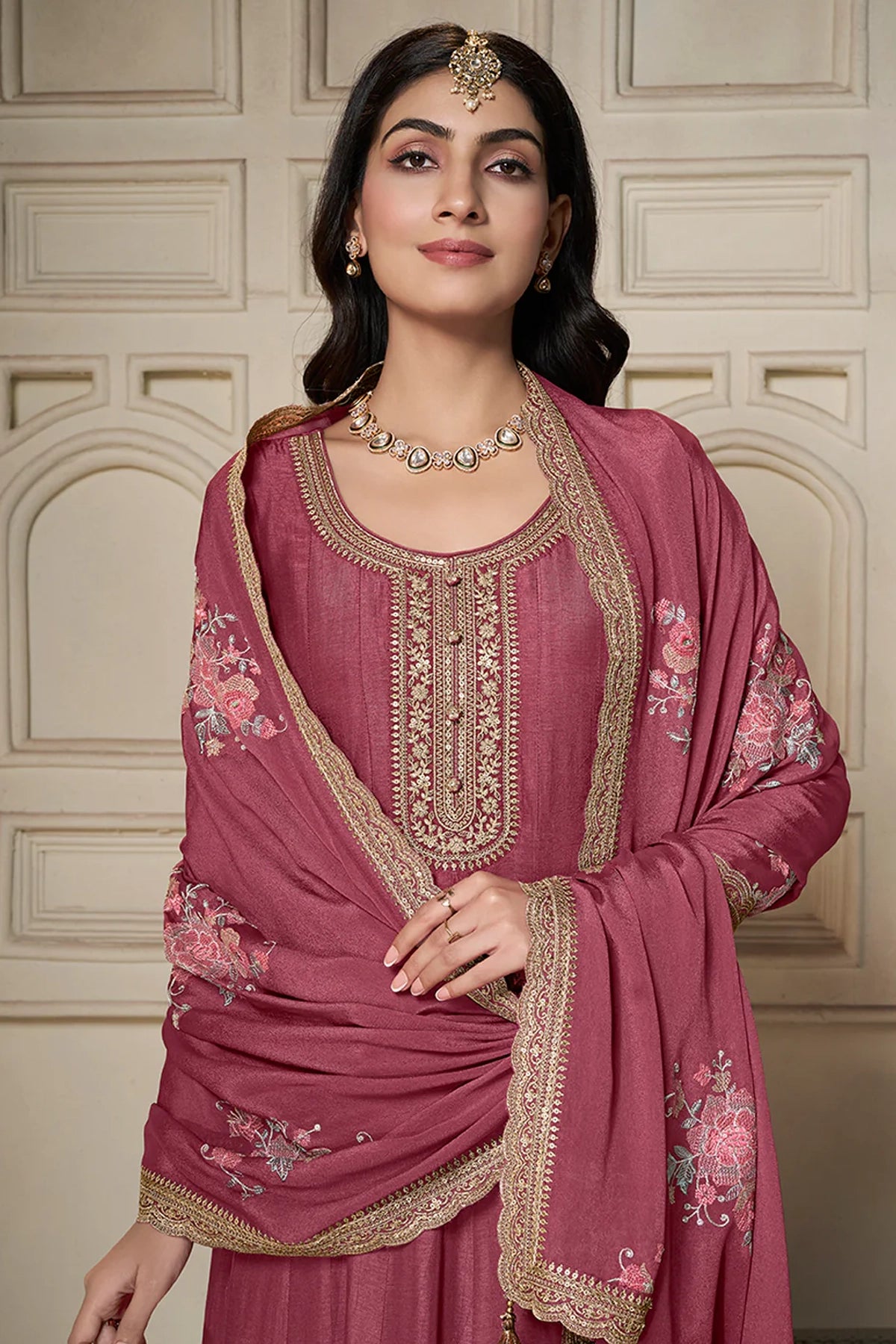 Woman wearing a traditional anarkali pink embroidered outfit with a matching dupatta against a beige wall.