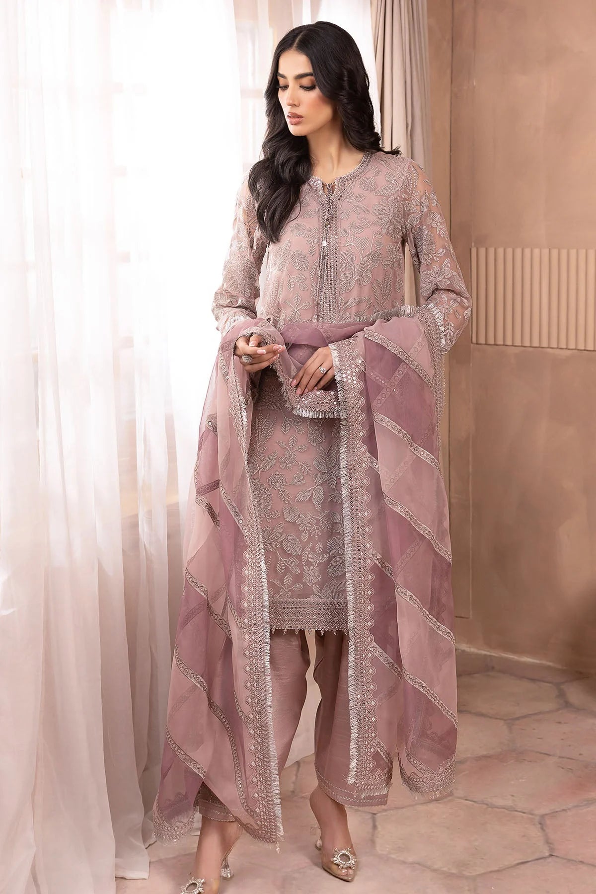 Woman wearing a pink embroidered traditional salwar suit outfit in a room with beige walls and white curtains.