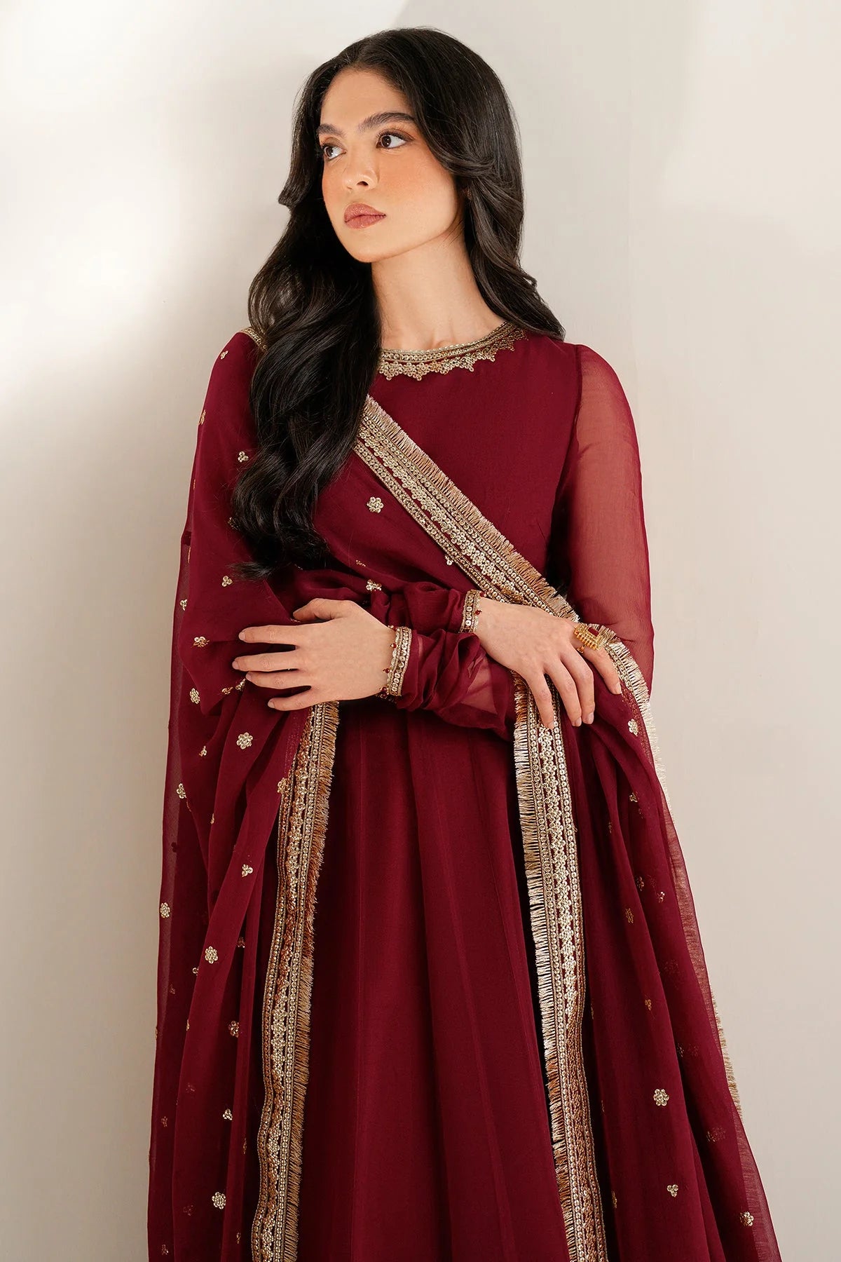 Woman wearing a long maroon anarkali dress with gold embroidery on a white background