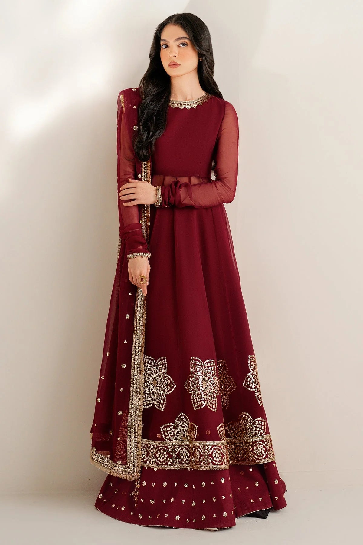 Woman wearing a long maroon anarkali dress with gold embroidery on a white background