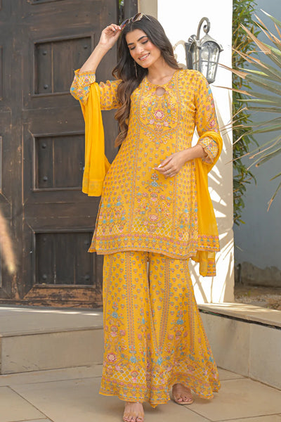 Woman wearing a yellow sharara suit embroidered traditional outfit standing in front of a wooden door.