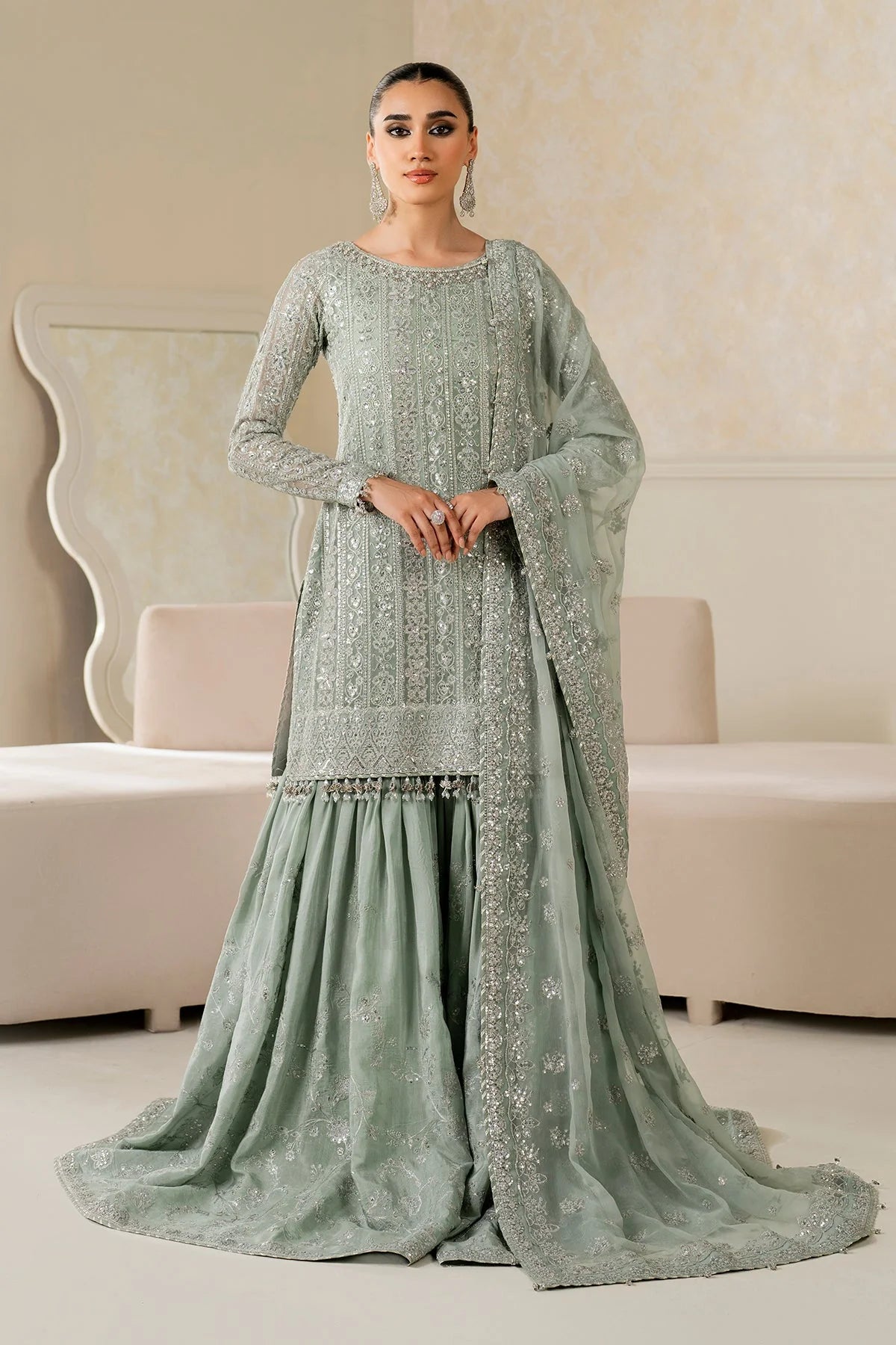 Woman wearing a light green traditional wedding sharara outfit with a matching dupatta in a room.