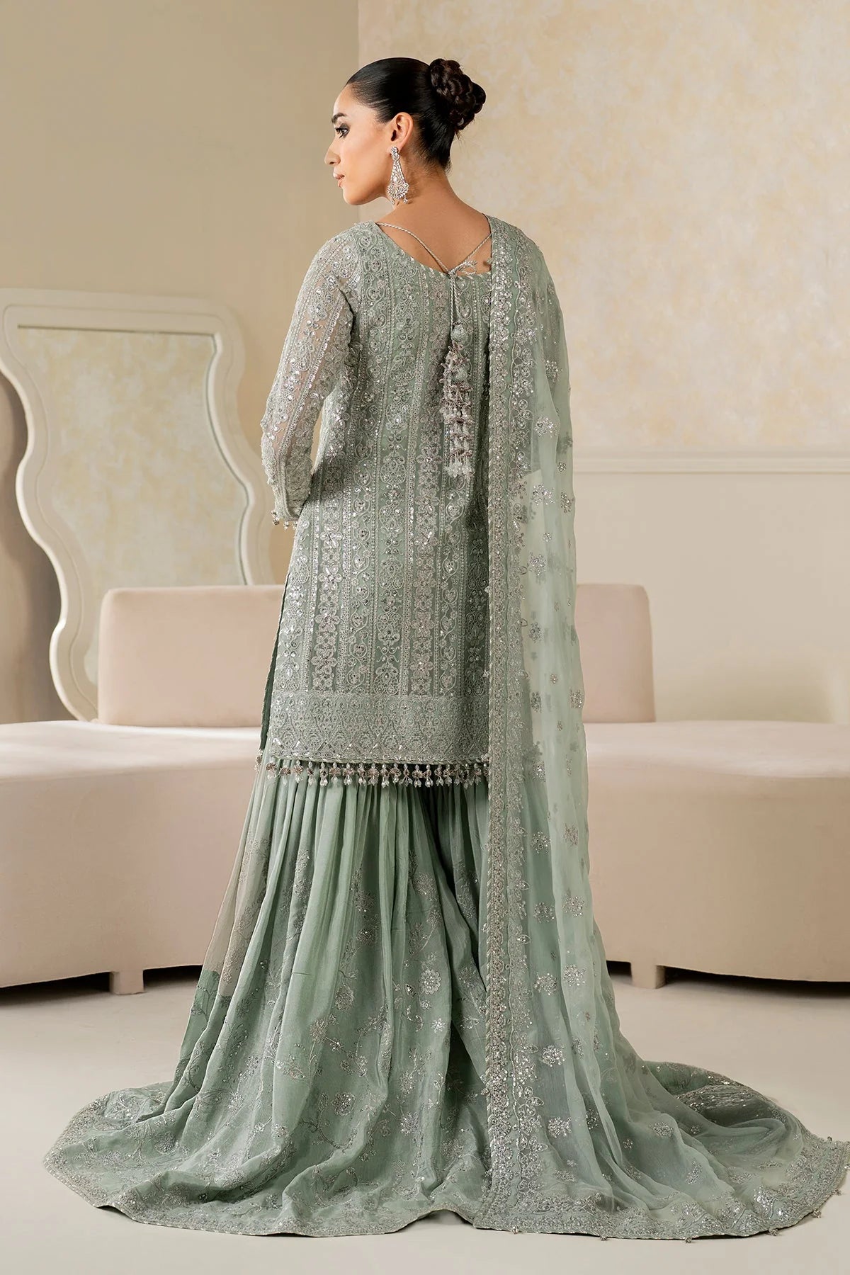 Woman wearing a light green traditional wedding sharara outfit with a matching dupatta in a room.