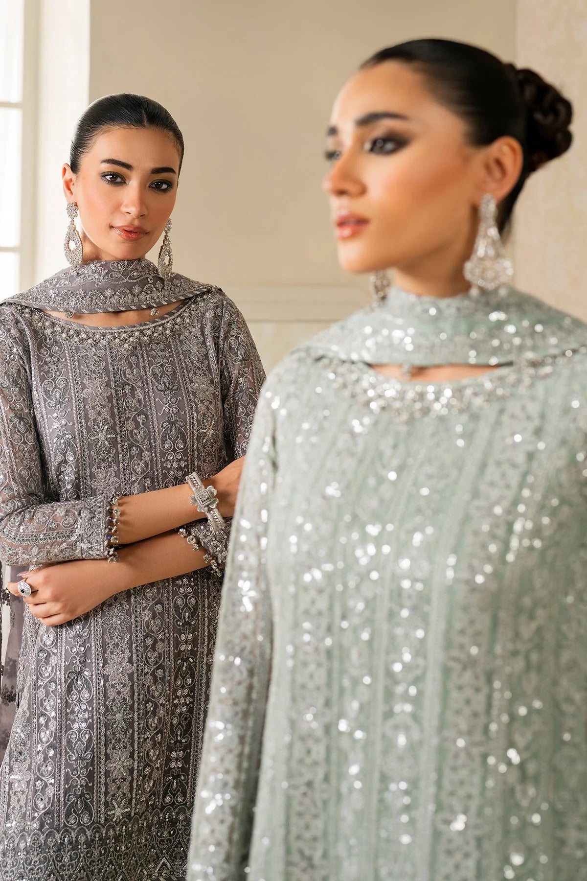 Two women wearing elegant dresses, one in gray and the other in silver, standing close to each other.