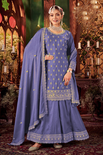 Woman in a traditional blue embroidered sharara style outfit with a matching dupatta in a decorative setting.