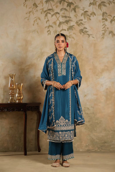 Woman wearing a blue embroidered traditional outfit standing against a beige wall.