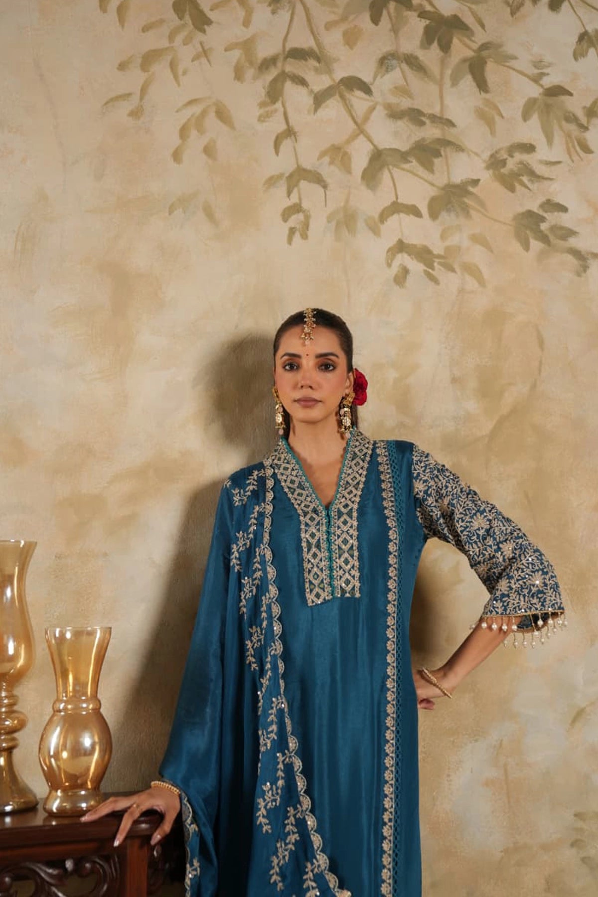 Woman in a blue embroidered outfit standing against a beige wall with decorative elements.