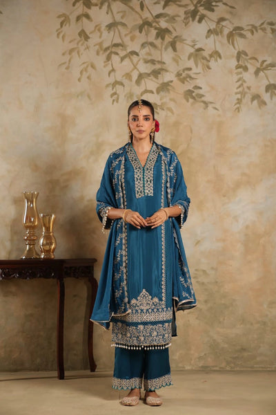 Woman in a blue embroidered traditional outfit standing against a beige wall with decorative elements.Woman in a blue embroidered traditional outfit standing against a beige wall with decorative elements.