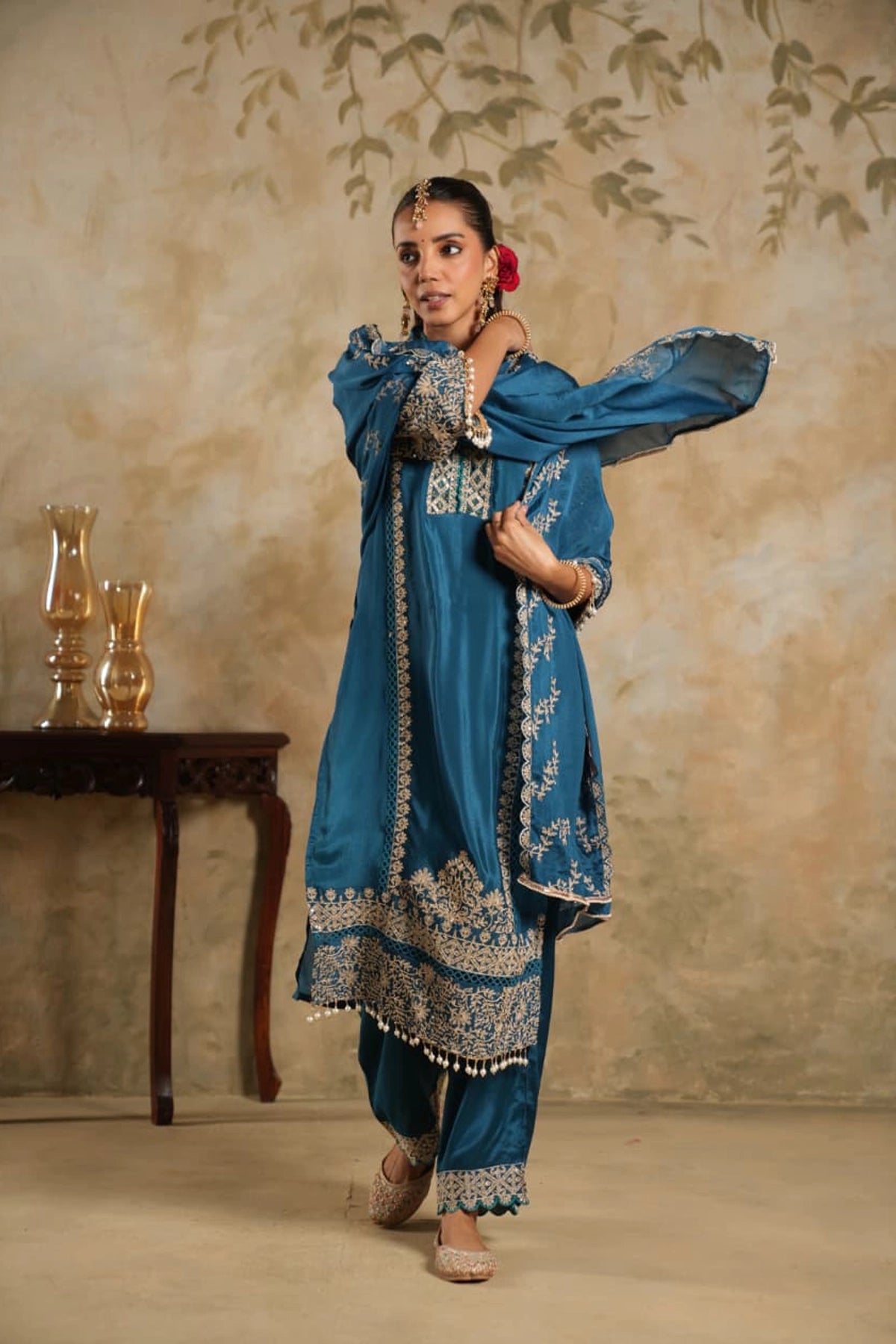Woman in a blue embroidered traditional outfit standing against a beige wall with decorative elements.
