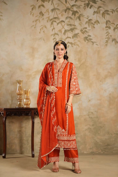 Woman wearing a traditional Dark Orange Chinon Silk Embroidered Kurta Set outfit with intricate patterns against a beige wall.
