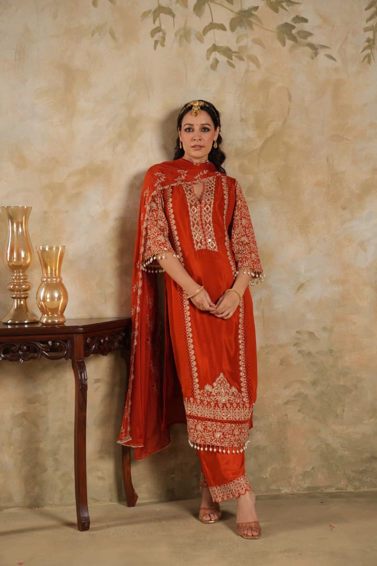 Woman wearing a traditional Dark Orange Chinon Silk Embroidered Kurta Set outfit with intricate patterns against a beige wall.