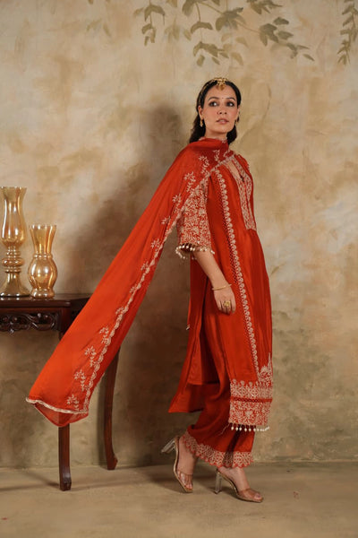 Woman wearing a traditional Dark Orange Chinon Silk Embroidered Kurta Set outfit with intricate patterns against a beige wall.Woman wearing a traditional Dark Orange Chinon Silk Embroidered Kurta Set outfit with intricate patterns against a beige wall.