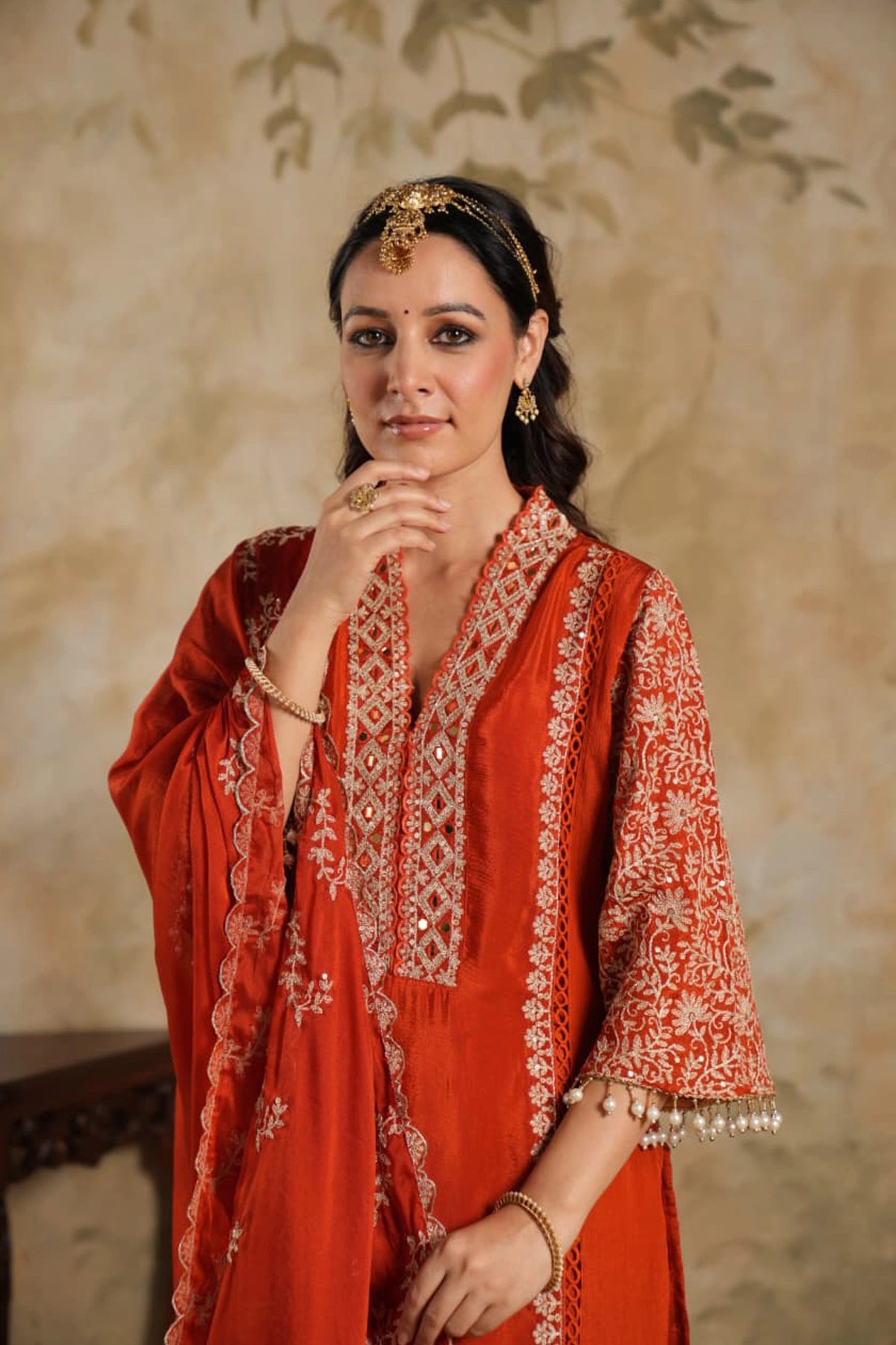Woman wearing a traditional Dark Orange Chinon Silk Embroidered Kurta Set outfit with intricate patterns against a beige wall.