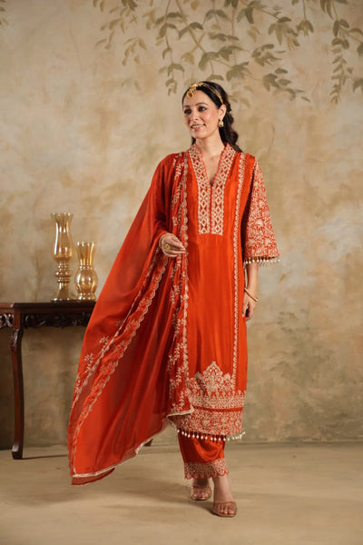 Woman wearing a traditional Dark Orange Chinon Silk Embroidered Kurta Set outfit with intricate patterns against a beige wall.Woman wearing a traditional Dark Orange Chinon Silk Embroidered Kurta Set outfit with intricate patterns against a beige wall.