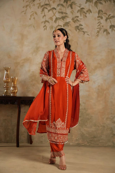 Woman wearing a traditional Dark Orange Chinon Silk Embroidered Kurta Set outfit with intricate patterns against a beige wall.Woman wearing a traditional Dark Orange Chinon Silk Embroidered Kurta Set outfit with intricate patterns against a beige wall.