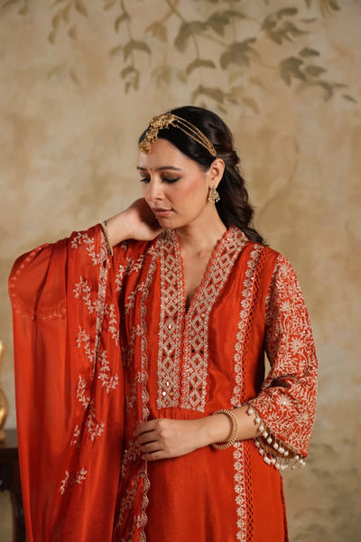 Woman wearing a traditional Dark Orange Chinon Silk Embroidered Kurta Set outfit with intricate patterns against a beige wall.Woman wearing a traditional Dark Orange Chinon Silk Embroidered Kurta Set outfit with intricate patterns against a beige wall.