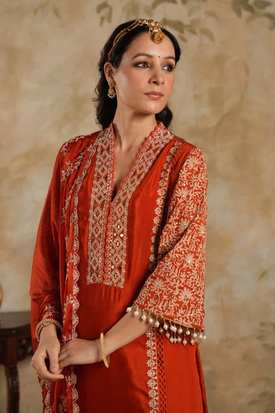 Woman wearing a traditional Dark Orange Chinon Silk Embroidered Kurta Set outfit with intricate patterns against a beige wall.Woman wearing a traditional Dark Orange Chinon Silk Embroidered Kurta Set outfit with intricate patterns against a beige wall.