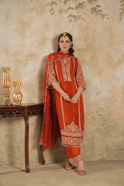 Woman wearing a traditional Dark Orange Chinon Silk Embroidered Kurta Set outfit with intricate patterns against a beige wall.Woman wearing a traditional Dark Orange Chinon Silk Embroidered Kurta Set outfit with intricate patterns against a beige wall.