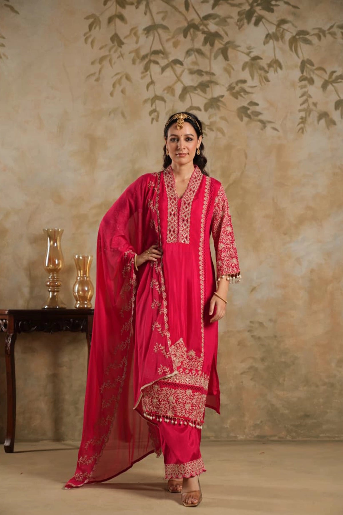 Woman in a rani pink traditional krta set outfit with gold embroidery against a beige wall.