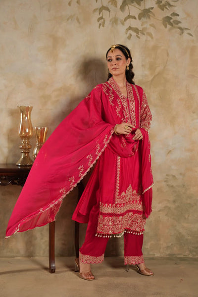 Woman in a rani pink traditional krta set outfit with gold embroidery against a beige wall.Woman in a rani pink traditional krta set outfit with gold embroidery against a beige wall.