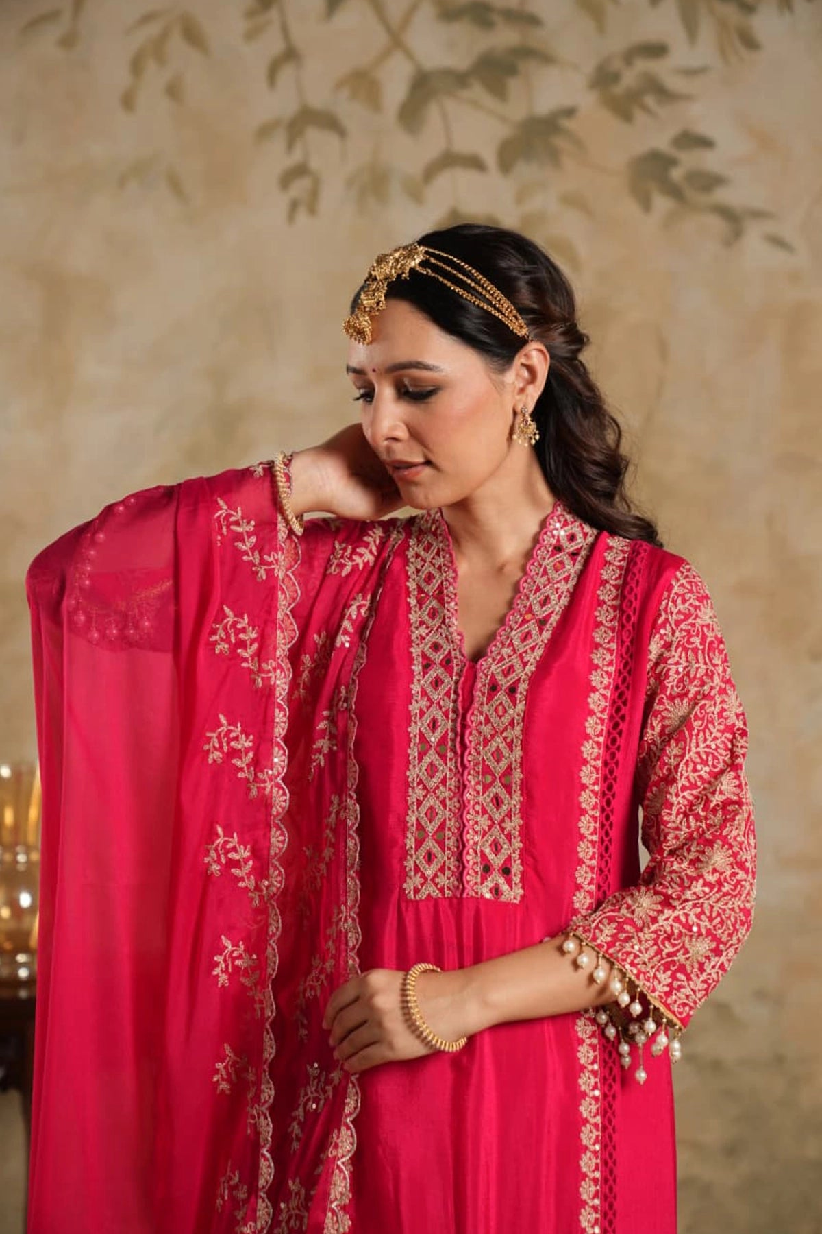 Woman in a rani pink traditional krta set outfit with gold embroidery against a beige wall.