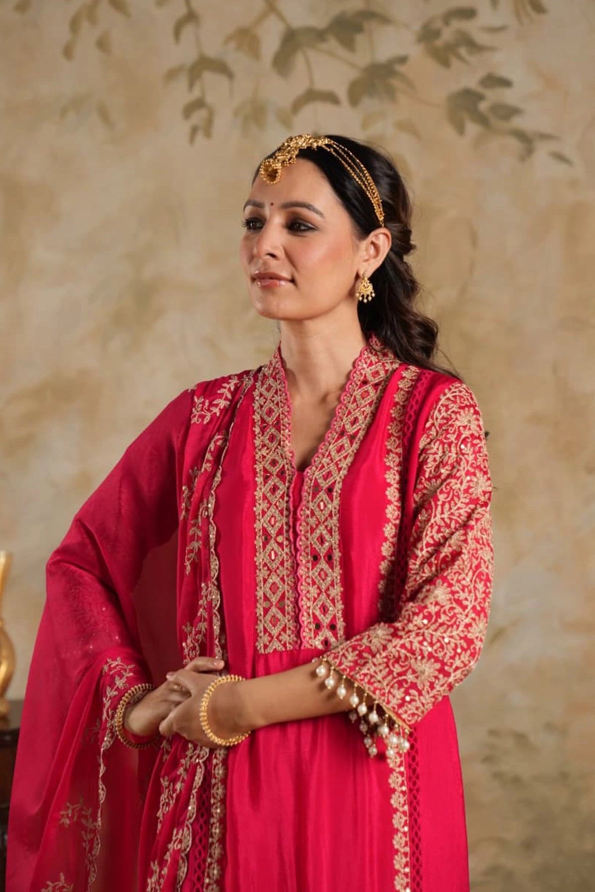 Woman in a rani pink traditional krta set outfit with gold embroidery against a beige wall.