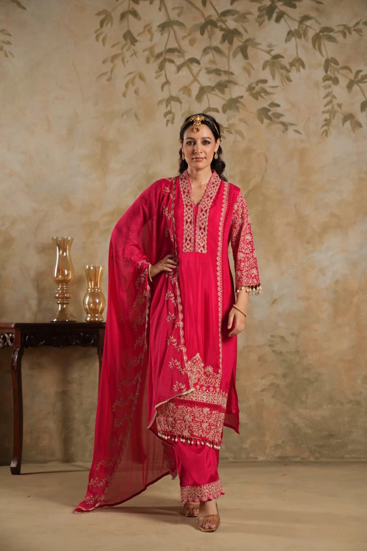 Woman in a rani pink traditional krta set outfit with gold embroidery against a beige wall.