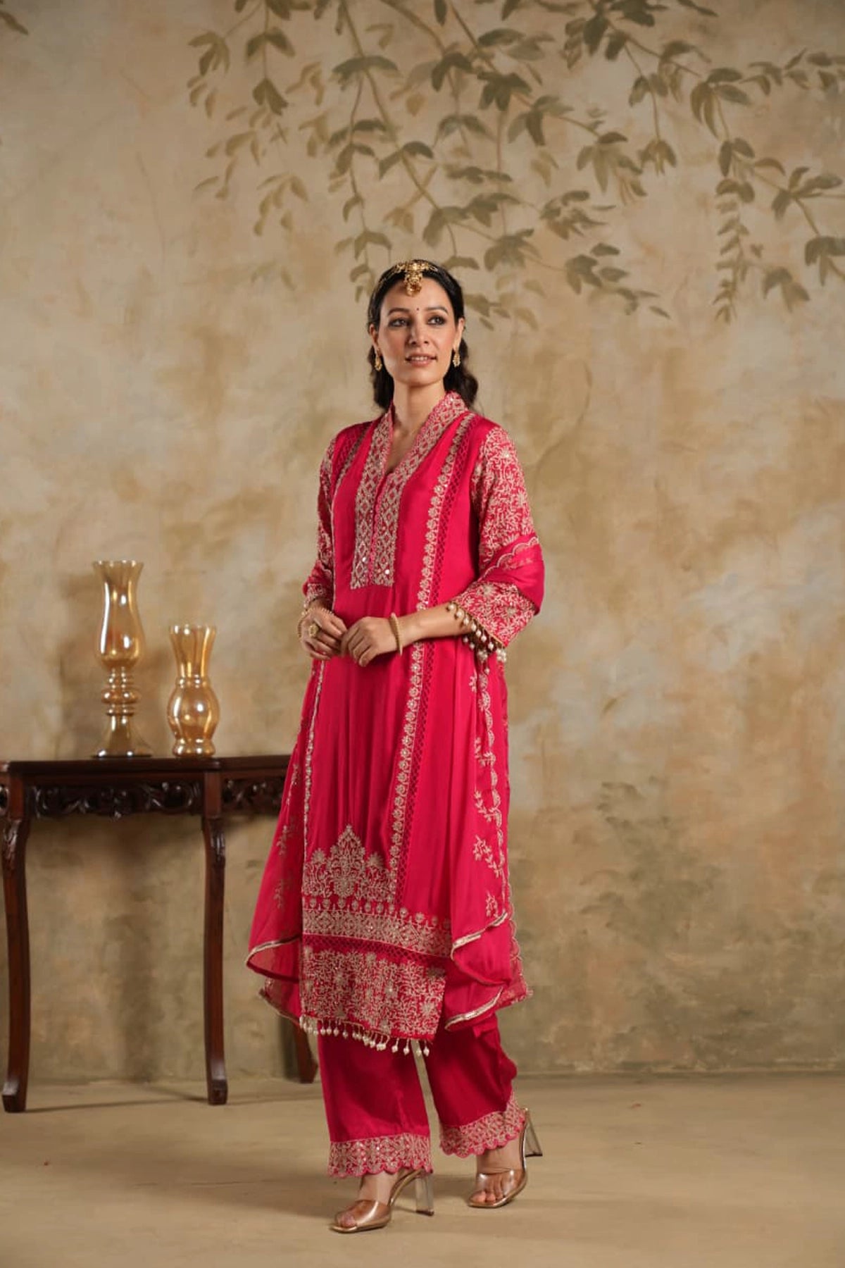 Woman in a rani pink traditional krta set outfit with gold embroidery against a beige wall.