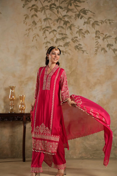 Woman in a rani pink traditional krta set outfit with gold embroidery against a beige wall.Woman in a rani pink traditional krta set outfit with gold embroidery against a beige wall.