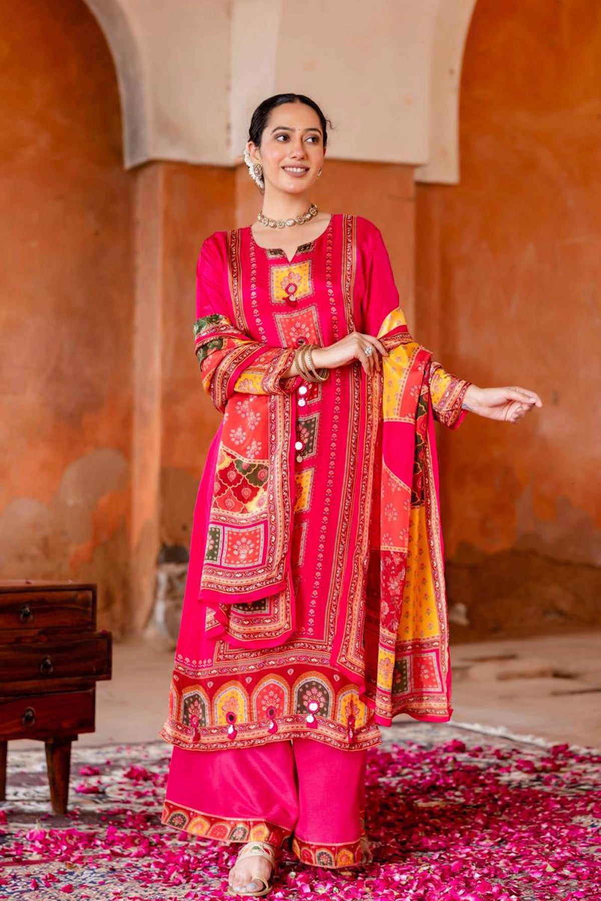 Woman in a vibrant pink traditional outfit with intricate patterns standing in a decorative setting.