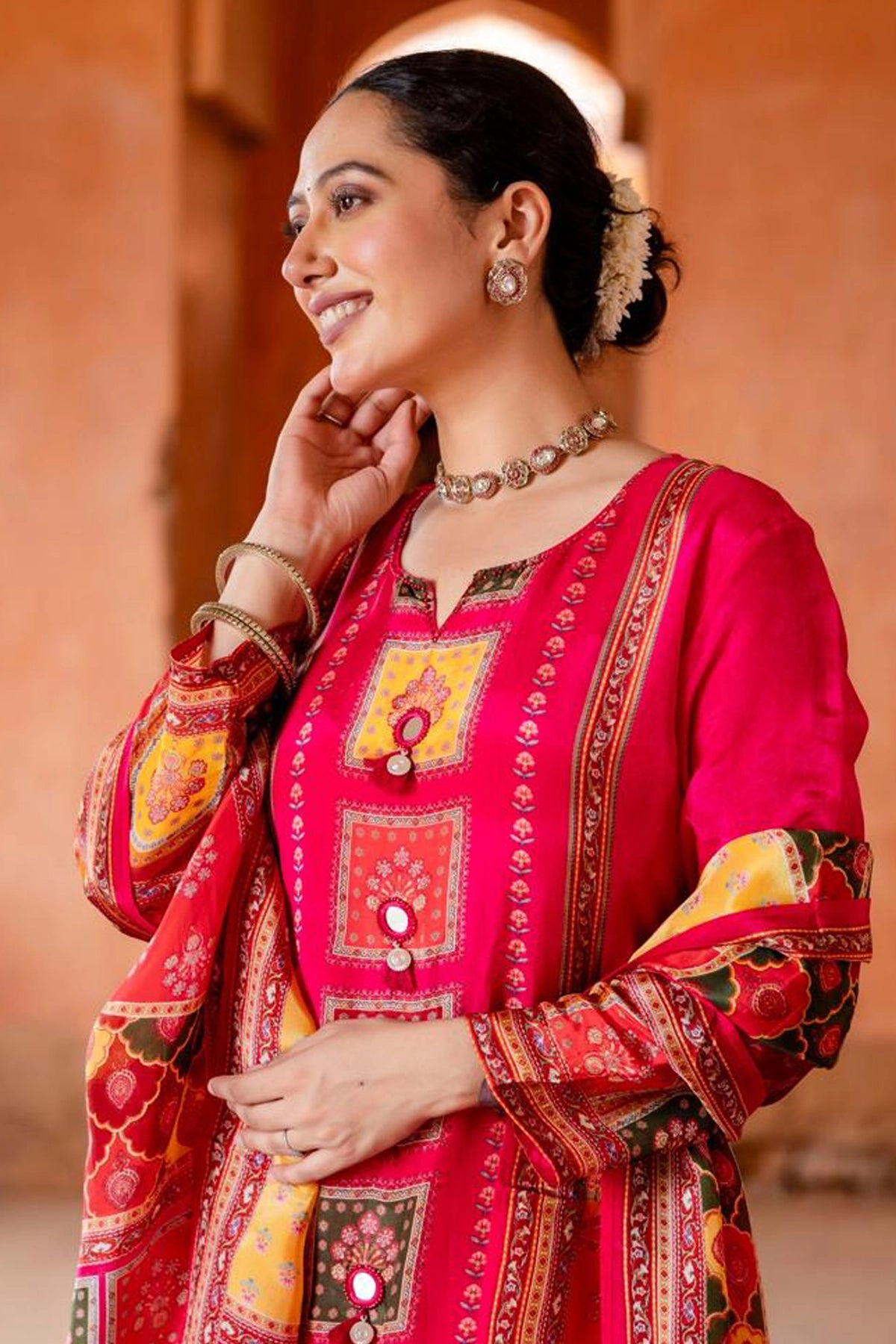 Woman wearing a traditional red and gold embroidered outfit with jewelry against an orange wall.