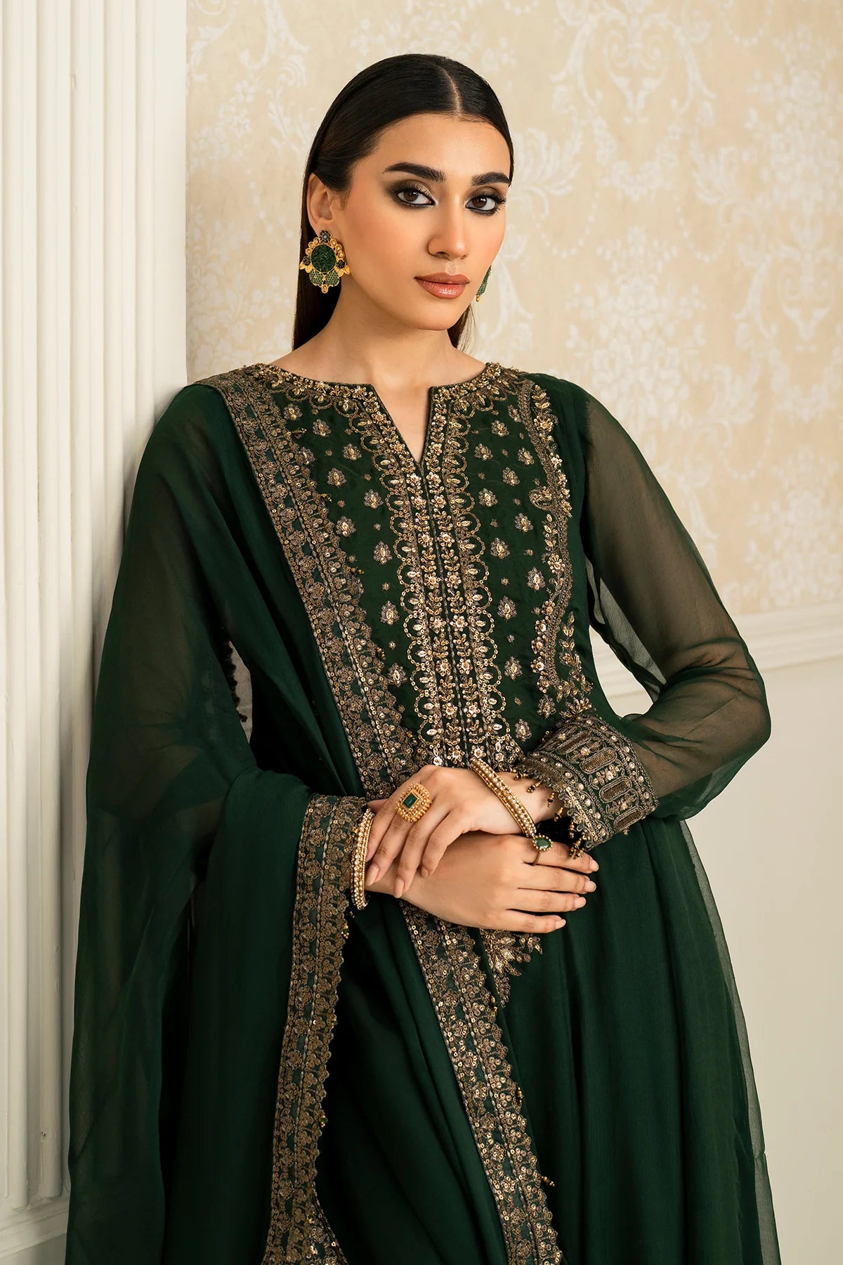 Woman wearing a dark green traditional anarkali outfit with intricate patterns against a beige wall.
