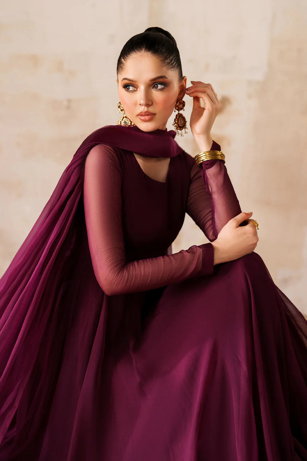 Woman wearing a burgundy chiffon anarkali dress with sheer sleeves against a beige wall.
