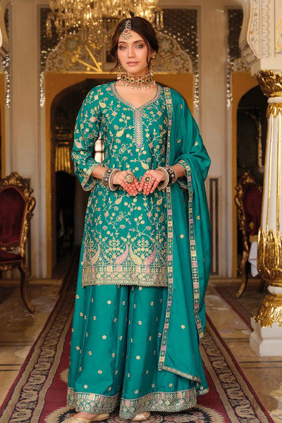 Woman in a teal embroidered sharara traditional outfit standing in an ornate room.
