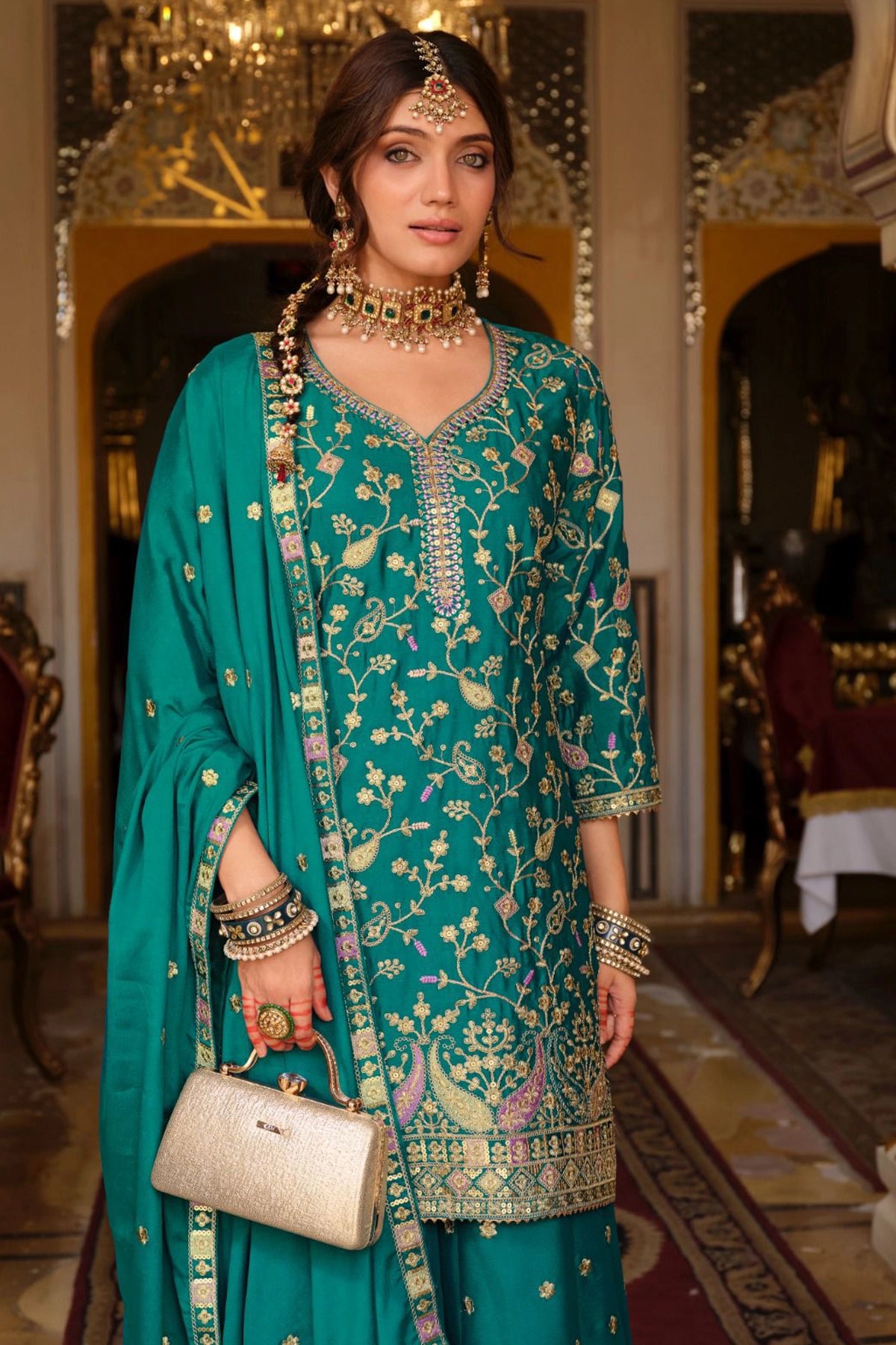 Woman in a teal traditional outfit with jewelry and a handbag in an ornate room.