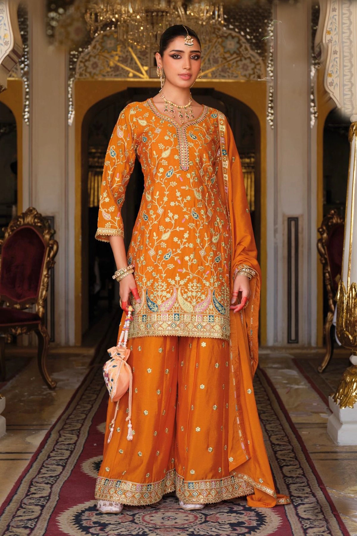 Woman in an orange traditional sharara outfit with intricate patterns in a decorated room.