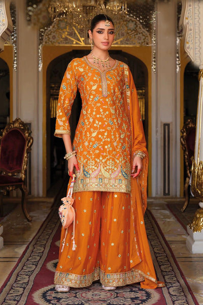 Woman in an orange traditional sharara outfit with intricate patterns in a decorated room.