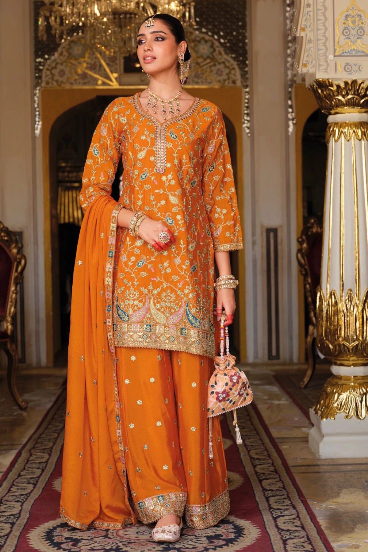 Woman in an orange traditional sharara outfit with intricate patterns in a decorated room.