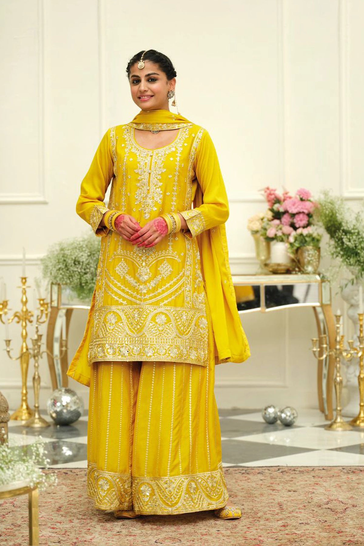 Woman in a yellow traditional sharara salwar suit outfit with intricate embroidery in an elegant indoor setting.