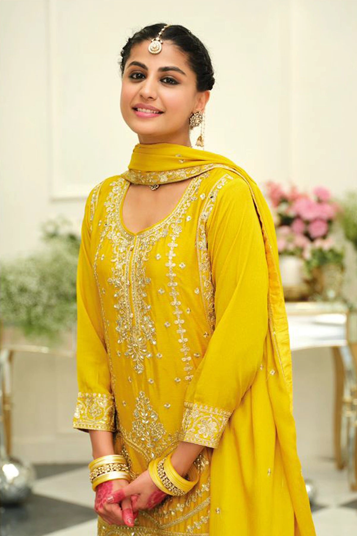Woman in a yellow traditional sharara salwar suit outfit with intricate embroidery in an elegant indoor setting.