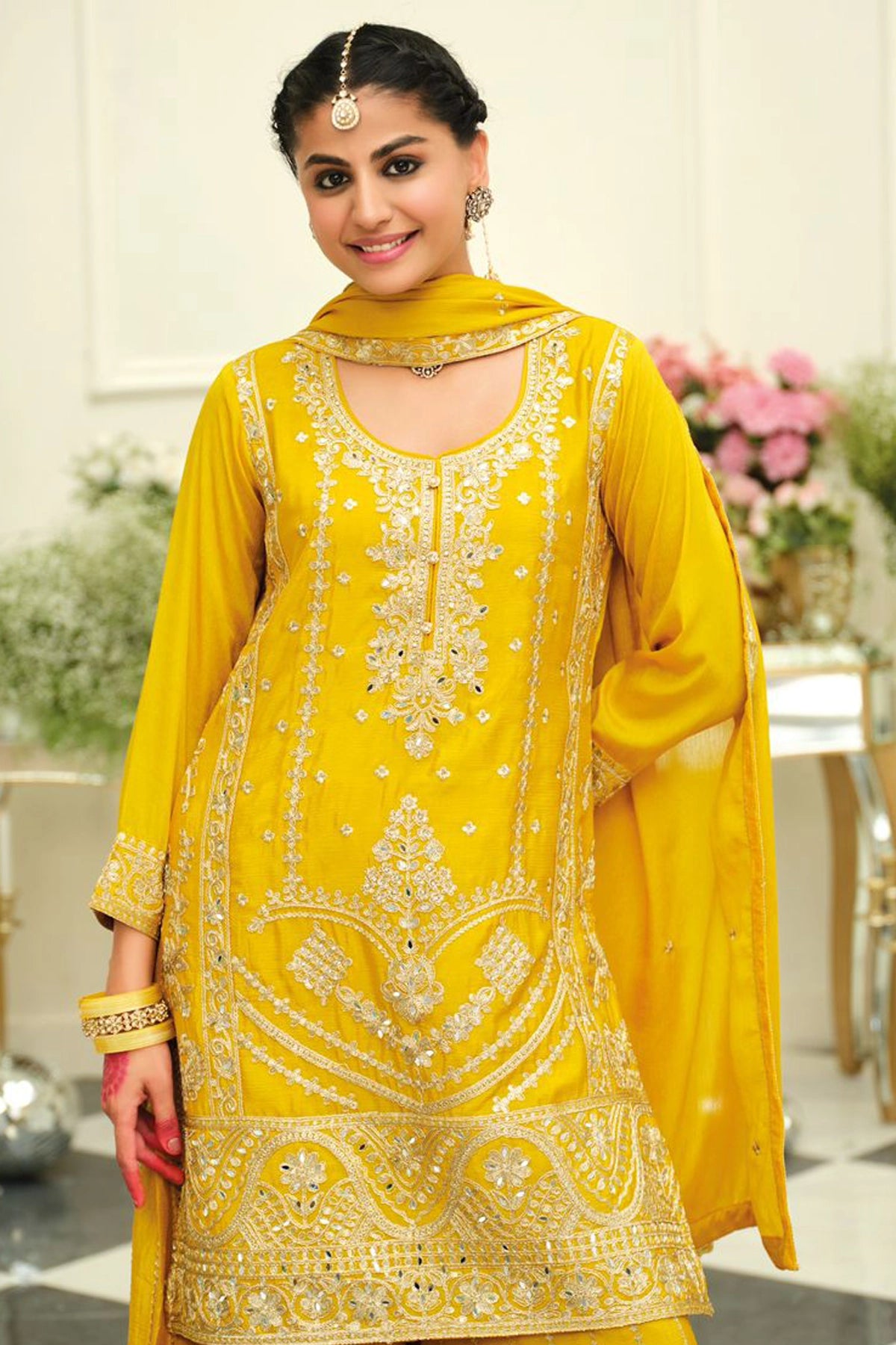 Woman in a yellow traditional sharara salwar suit outfit with intricate embroidery in an elegant indoor setting.