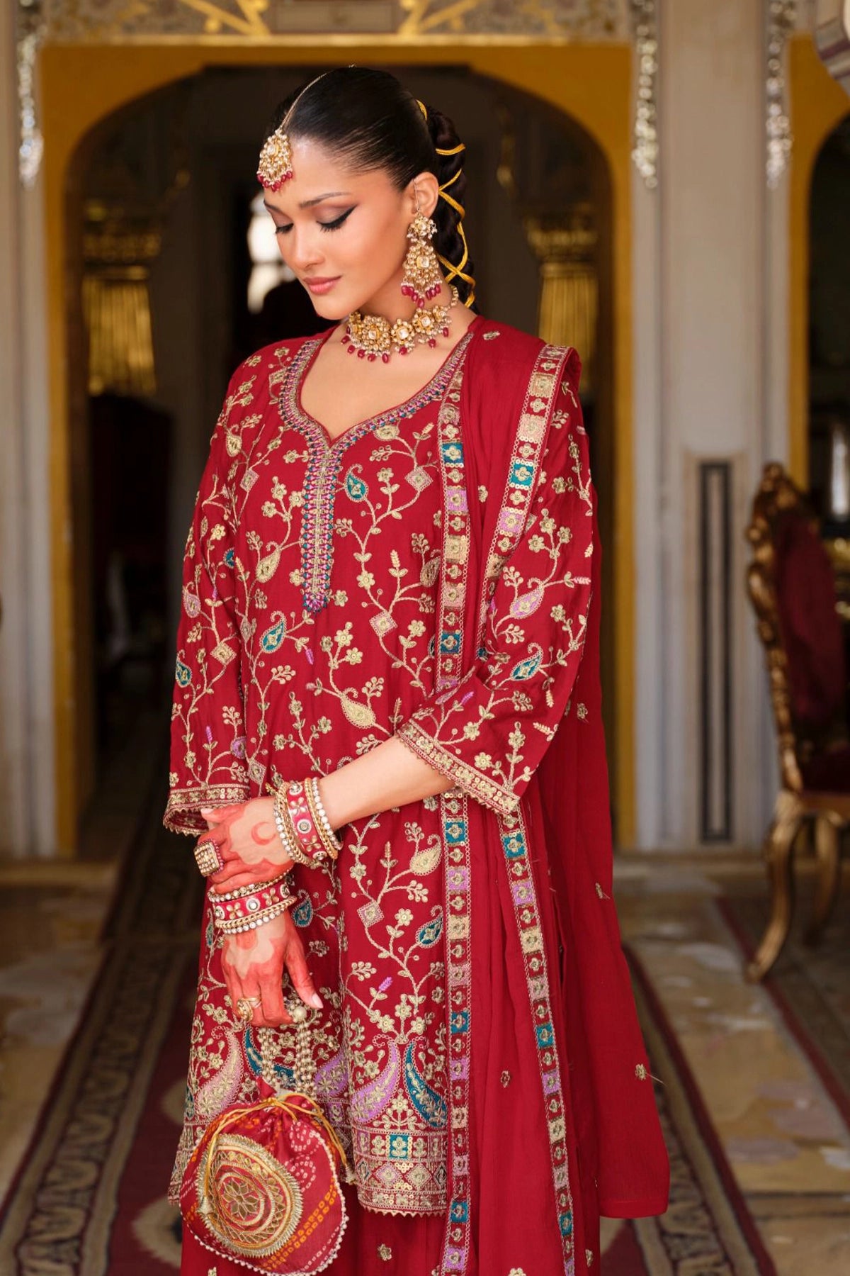 Woman in a traditional red embroidered sharara outfit standing in an ornate room.