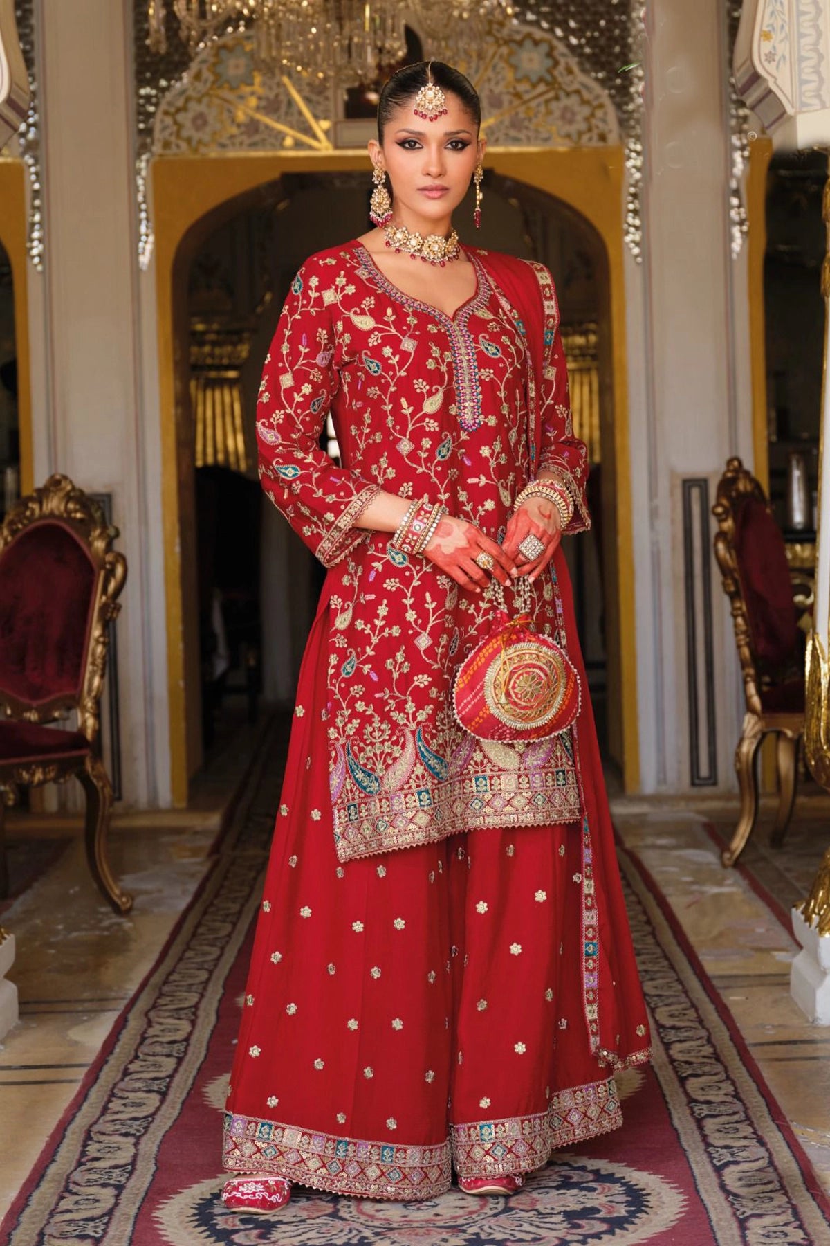 Woman in a traditional red embroidered sharara outfit standing in an ornate room.