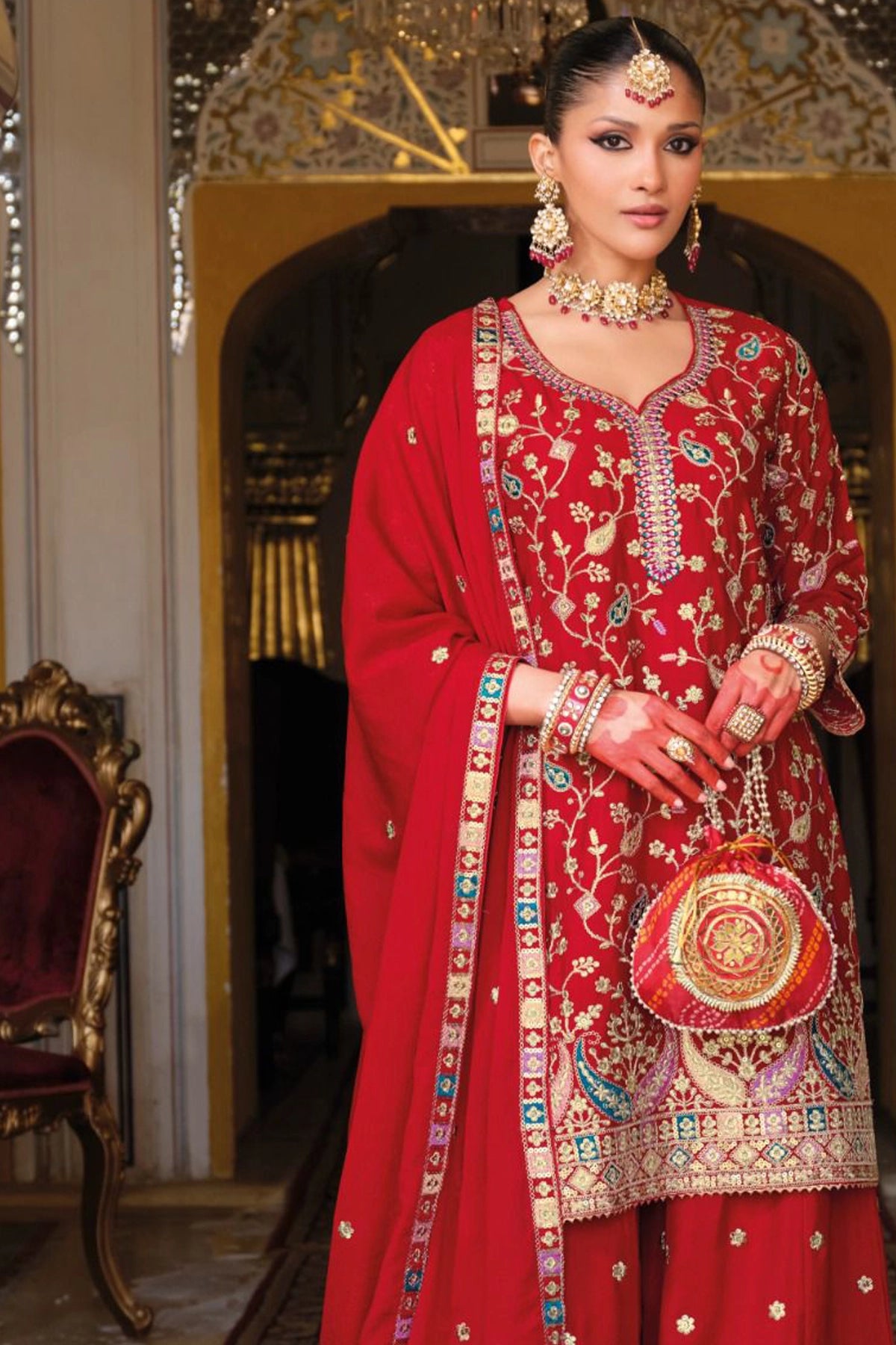 Woman in a traditional red embroidered sharara outfit standing in an ornate room.