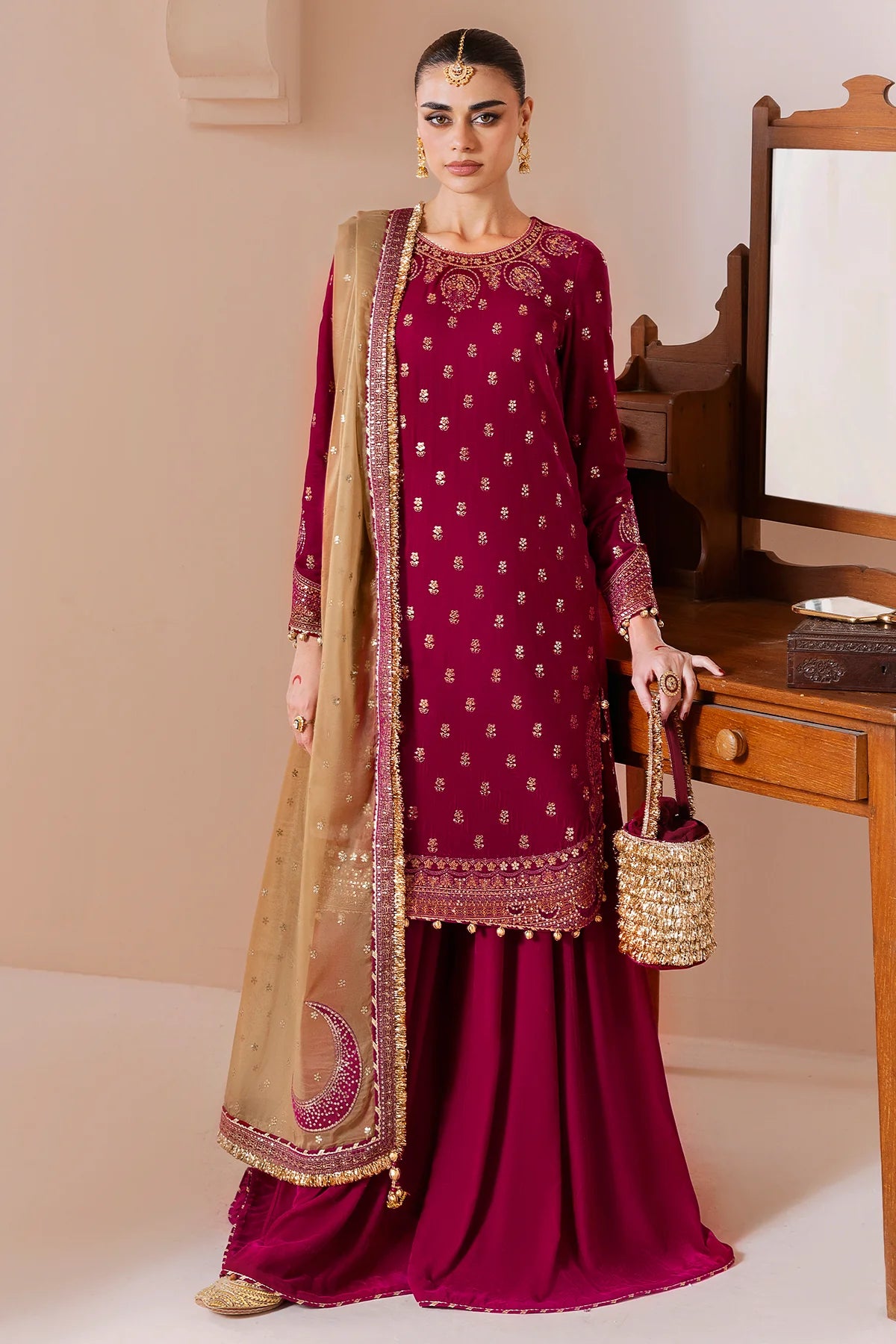 Woman in a maroon traditional velvet sharara outfit with gold embroidery, holding a matching bag, against a neutral background.