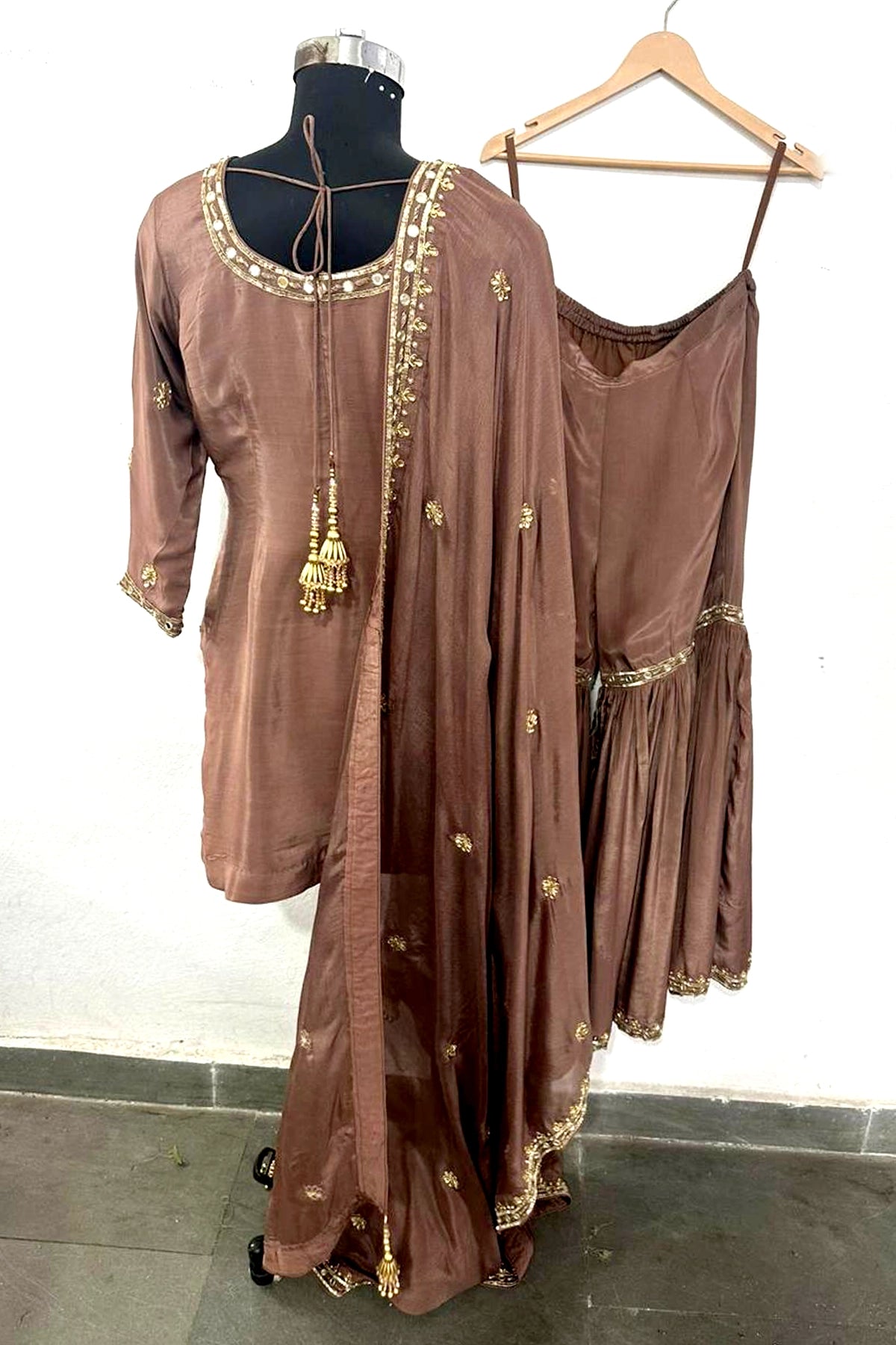 Brown traditional outfit with gold embroidery on a mannequin and hanger against a white wall.