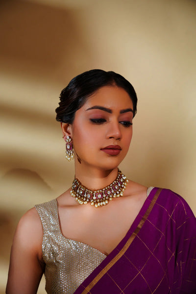 Woman wearing a purple saree with gold border and jewelry against a beige background