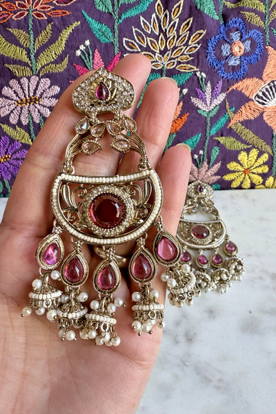 Ornate gold earrings with purple gemstones held in a hand against a colorful embroidered fabric background.Ornate gold earrings with purple gemstones held in a hand against a colorful embroidered fabric background.