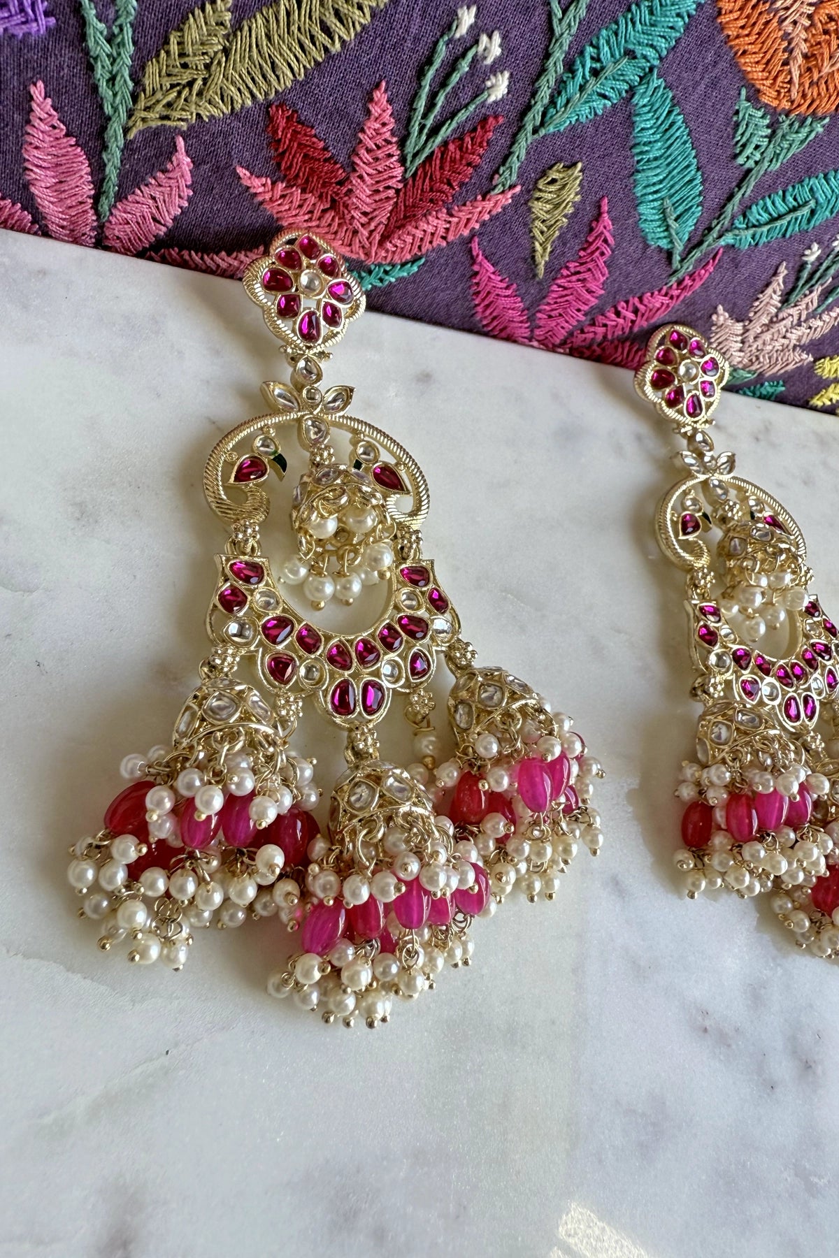 Jumka earrings with pink and white pearls, and stones on a marble surface with an embroidered fabric background.