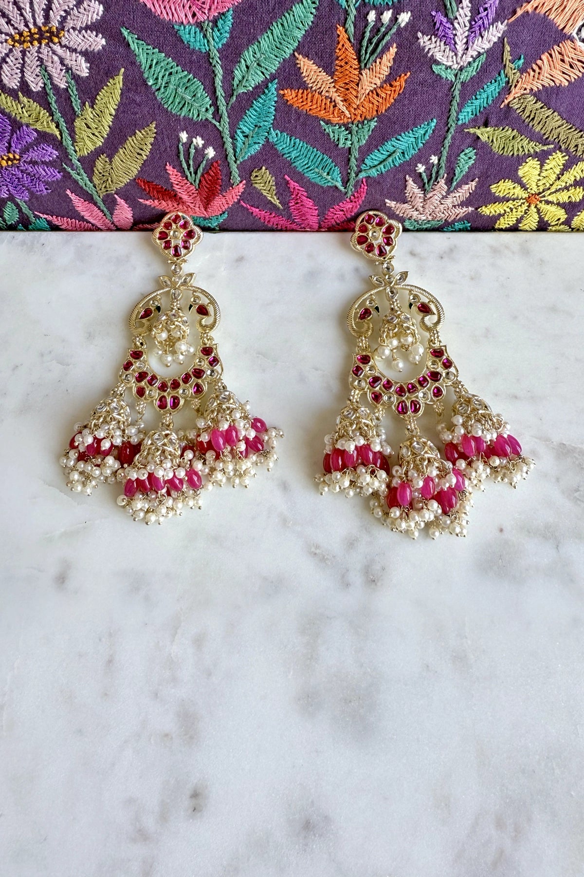 Jumka earrings with pink and gold details on a marble surface with a colorful fabric background.